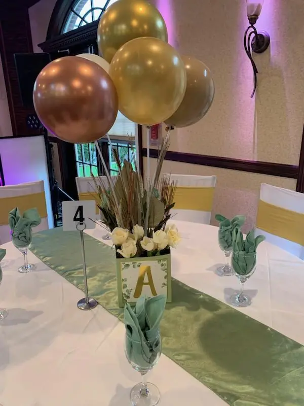 Table number centerpiece with gold balloons, tall dried grasses and white flowers on a sage runner
