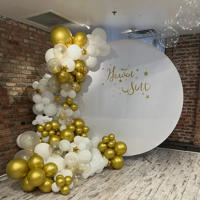 Gold, white and gold-confetti balloon garland climbing a round white backdrop against exposed brick