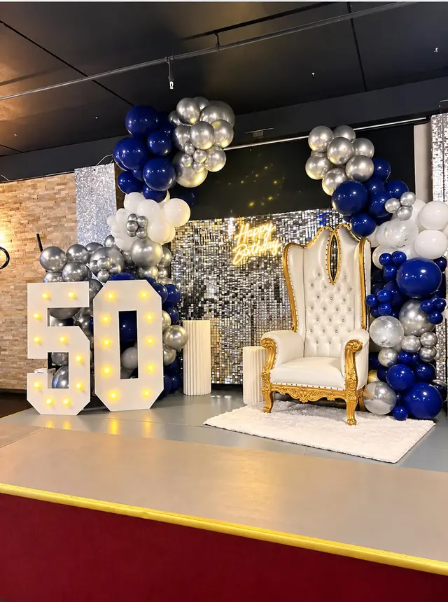 Light-up 50 marquee numbers beside a white throne chair under navy, silver and white balloons on a stage