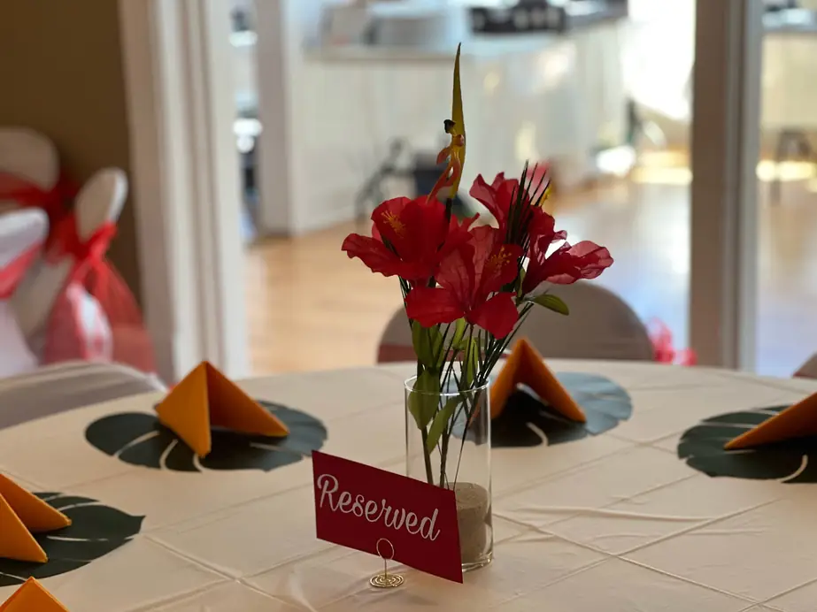 Round table with white linen, folded orange napkins and red lilies beside a Reserved sign